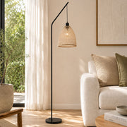 A modern black floor lamp with a curved arm and a woven natural-fiber conical shade stands in a sunlit living room beside a cream sofa and large window - Lighting.co.za