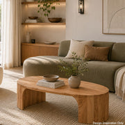 A modern living room scene featuring a low, rounded wooden coffee table in the foreground, with a sage green sectional sofa and neutral throw pillows in the background. The room is styled with warm lighting, a wooden sideboard, and shelves - Lighting.co.za