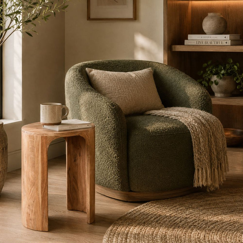 An olive green bouclé armchair in a warm, styled room setting with a beige throw pillow, a beige throw blanket, a small wooden side table with a mug, and a wooden bookshelf in the background - Lighting.co.za