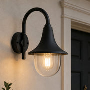 A black outdoor wall sconce with a curved arm and clear PVC shade, mounted on a beige stucco wall next to a dark door. Perfect for Coastal areas - Lighting.co.za