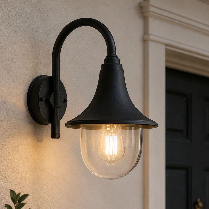 A black outdoor wall sconce with a curved arm and clear PVC shade, mounted on a beige stucco wall next to a dark door. Perfect for Coastal areas - Lighting.co.za