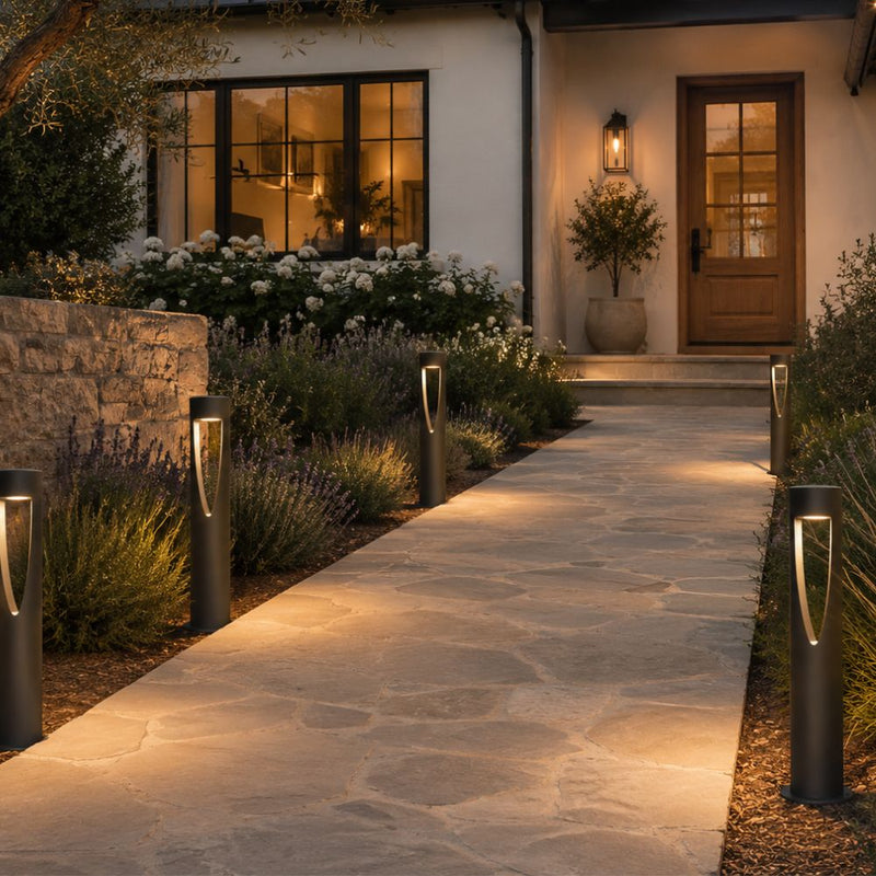 Modern outdoor pathway lights installed along a stone walkway leading to a house entrance, with warm light glowing from the sleek black bollard posts in a landscaped garden setting - Lighting.co.za