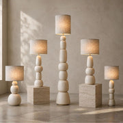 Five modern table lamps with stacked spherical bases and cylindrical fabric shades are arranged on matching stone pedestals against a textured beige wall. The lamps are shown in five different heights, all lit to cast a warm glow - Lighting.co.za