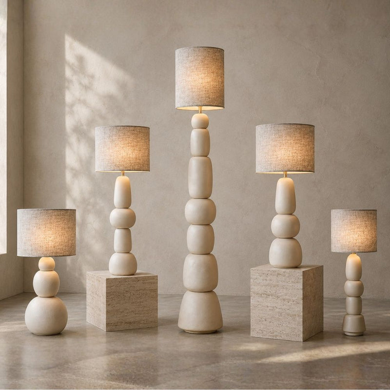 Five modern table lamps with stacked spherical bases and cylindrical fabric shades are arranged on matching stone pedestals against a textured beige wall. The lamps are shown in five different heights, all lit to cast a warm glow - Lighting.co.za