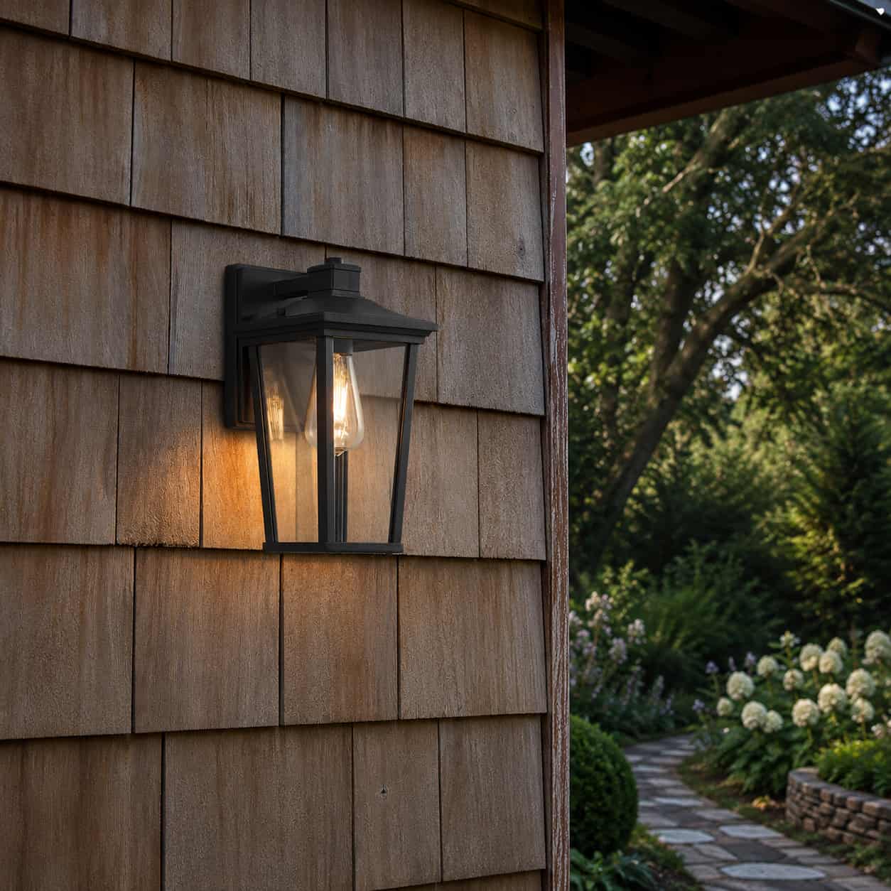 A black outdoor wall lantern light fixture mounted on a brown wood shingle house exterior, casting a warm glow onto the siding, with a garden path and trees visible in the background - Lighting.co.za