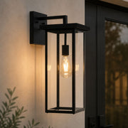 A black outdoor wall sconce with a clear glass lantern-style shade and a visible warm Edison bulb, mounted on a beige stucco wall beside a dark door - Lighting.co.za