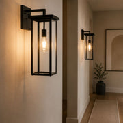 A black metal outdoor wall sconce with a rectangular frame and visible Edison bulb, mounted on a beige wall in a hallway. A second matching sconce appears further down the hall, with a plant and framed wall art in the background - Lighting.co.za