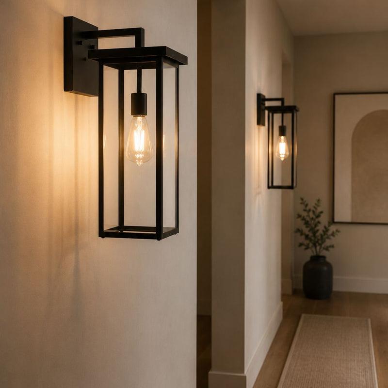 A black metal outdoor wall sconce with a rectangular frame and visible Edison bulb, mounted on a beige wall in a hallway. A second matching sconce appears further down the hall, with a plant and framed wall art in the background - Lighting.co.za