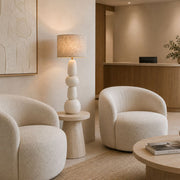 A modern, warm-toned interior featuring two rounded cream bouclé armchairs, a sculptural stacked ceramic table lamp with a textured beige shade, and light wood furniture in a minimalist lounge or reception area - Lighting.co.za