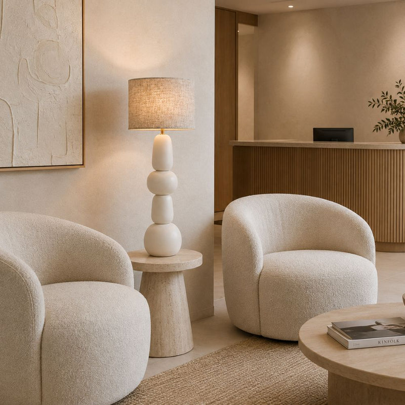A modern, warm-toned interior featuring two rounded cream bouclé armchairs, a sculptural stacked ceramic table lamp with a textured beige shade, and light wood furniture in a minimalist lounge or reception area - Lighting.co.za