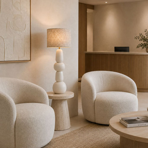A modern, warm-toned interior featuring two rounded cream bouclé armchairs, a sculptural stacked ceramic table lamp with a textured beige shade, and light wood furniture in a minimalist lounge or reception area - Lighting.co.za