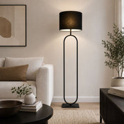 A modern black floor lamp with a cylindrical drum shade and a tall, curved metal frame stands in a styled living room next to a light beige sofa and a dark wood side table - Lighting.co.za