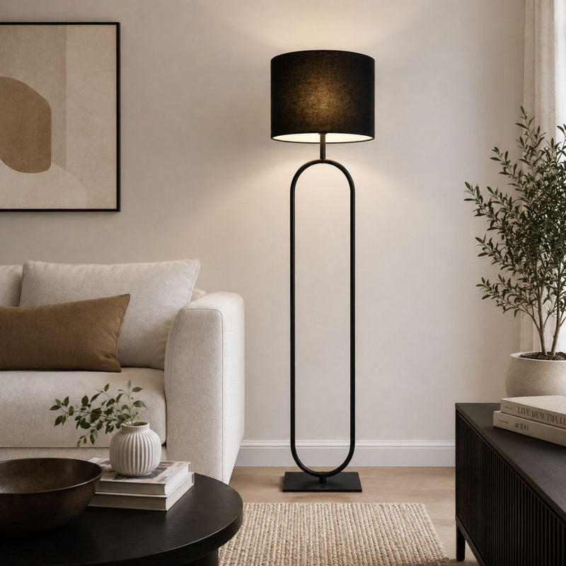 A modern black floor lamp with a cylindrical drum shade and a tall, curved metal frame stands in a styled living room next to a light beige sofa and a dark wood side table - Lighting.co.za