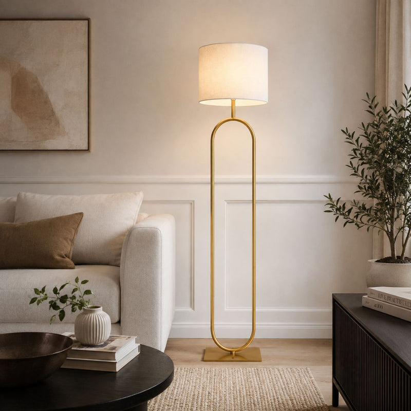 A modern floor lamp with a tall cylindrical white shade and a slim gold metal frame, standing in a styled living room next to a white sofa and a dark wood side table - Lighting.co.za