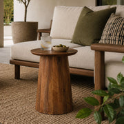 A round wooden side table with a flared pedestal base, styled in a modern living room next to a cream sofa with green and patterned pillows.