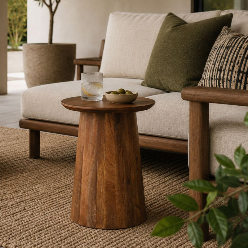 A round wooden side table with a flared pedestal base, styled in a modern living room next to a cream sofa with green and patterned pillows.