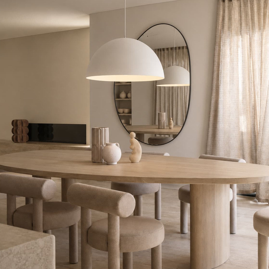 A modern dining room scene featuring a light wood oval dining table with upholstered chairs, a large white dome pendant light, and a round wall mirror reflecting the space.