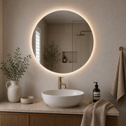 A round backlit LED bathroom mirror mounted on a beige wall above a marble countertop with a white vessel sink, gold faucet, and wooden vanity. Decorative plants, a ceramic vase, and a hanging towel are visible in the styled bathroom setting - Lighting.co.za