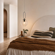 A modern bedroom scene featuring a clear glass globe pendant light hanging above a bed with a textured brown throw blanket, striped pillows, and a large arched headboard against warm beige walls - Lighting.co.za