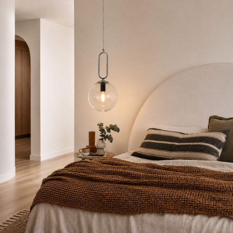 A modern bedroom scene featuring a clear glass globe pendant light hanging above a bed with a textured brown throw blanket, striped pillows, and a large arched headboard against warm beige walls - Lighting.co.za