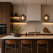 A modern kitchen interior with dark wood cabinetry, a light stone countertop, two hanging glass pendant lights with gold-toned loops, a white range hood, and three wooden bar stools at the island - Lighting.co.za