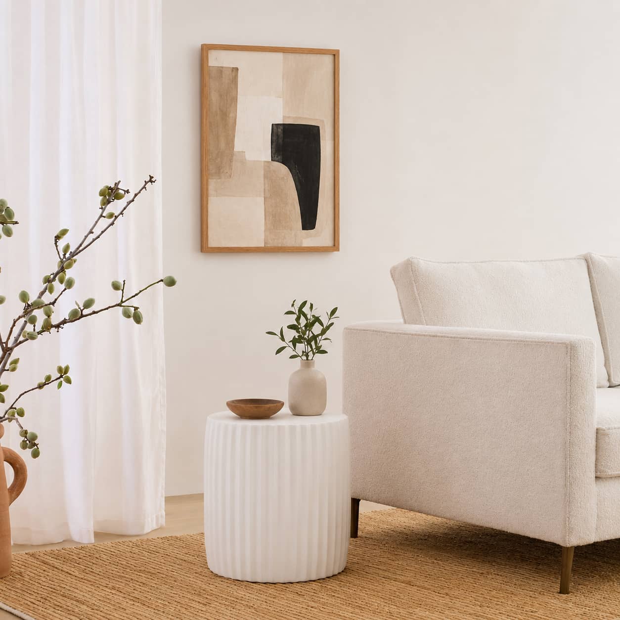 A styled living room corner featuring a white ribbed side table with a small vase and bowl, next to a white armchair. A framed abstract geometric print hangs on the wall above, with a curtain, woven rug, and potted plant in the background.