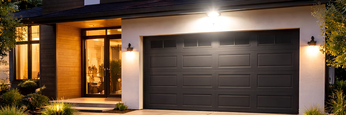 A modern single-car garage door in dark charcoal gray, installed on a white and wood exterior house with warm outdoor wall lights turned on. Outdoor Lighting Buying Guide - Lighting.co.za