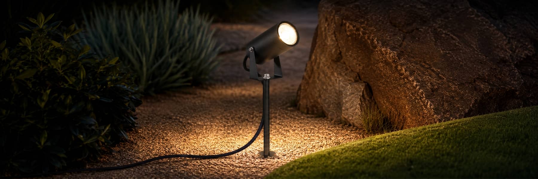 A black ground-mounted spotlight landscape light illuminating a path between rocks and plants at night. Outdoor Lighting Buying Guide - Lighting.co.za