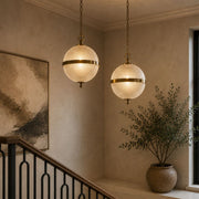 Two ribbed glass and antique brass globe pendant lights hang at different heights over a modern stairwell landing with warm neutral walls, a timber handrail, large artwork and indoor plant - Lighting.co.za