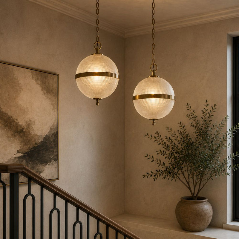 Two ribbed glass and antique brass globe pendant lights hang at different heights over a modern stairwell landing with warm neutral walls, a timber handrail, large artwork and indoor plant - Lighting.co.za