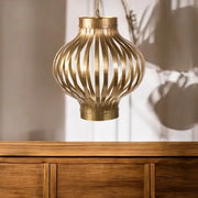 A hanging pendant light with a metallic gold finish and a ribbed, openwork lantern-like shade, shown above a wooden cabinet against a softly lit wall - Lighting.co.za