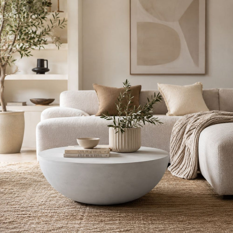 A modern round light grey coffee table in a neutral-toned living room setting, styled with a small potted olive branch and books, in front of a beige sectional sofa - Lighting.co.za