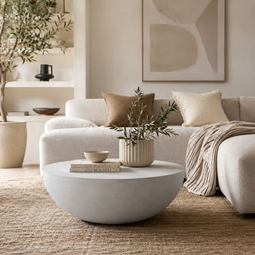 A modern round light grey coffee table in a neutral-toned living room setting, styled with a small potted olive branch and books, in front of a beige sectional sofa - Lighting.co.za