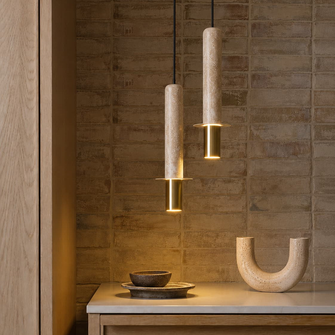 Two modern pendant lights with cylindrical beige stone or wood shades and gold-toned metal accents, hanging over a wooden console table in a warm interior setting.