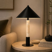 A modern table lamp with a matte black conical shade and base, accented by stacked cream-colored stone rings on the stem, shown lit on a round stone side table in a cozy living room setting - Lighting.co.za