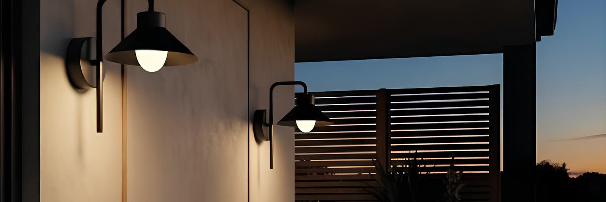 Two modern black outdoor wall sconces with conical shades and glowing warm-white bulbs mounted on a beige exterior wall at dusk, with a slatted fence and twilight sky in the background. Outdoor Lighting Buying Guide - Lighting.co.za