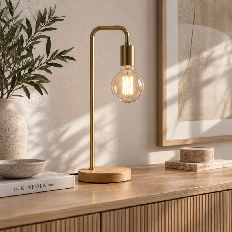 A gold table lamp with a curved metal stand and a clear glass bulb, lit with a warm glow, placed on a wooden console table in a styled interior setting - Lighting.co.za