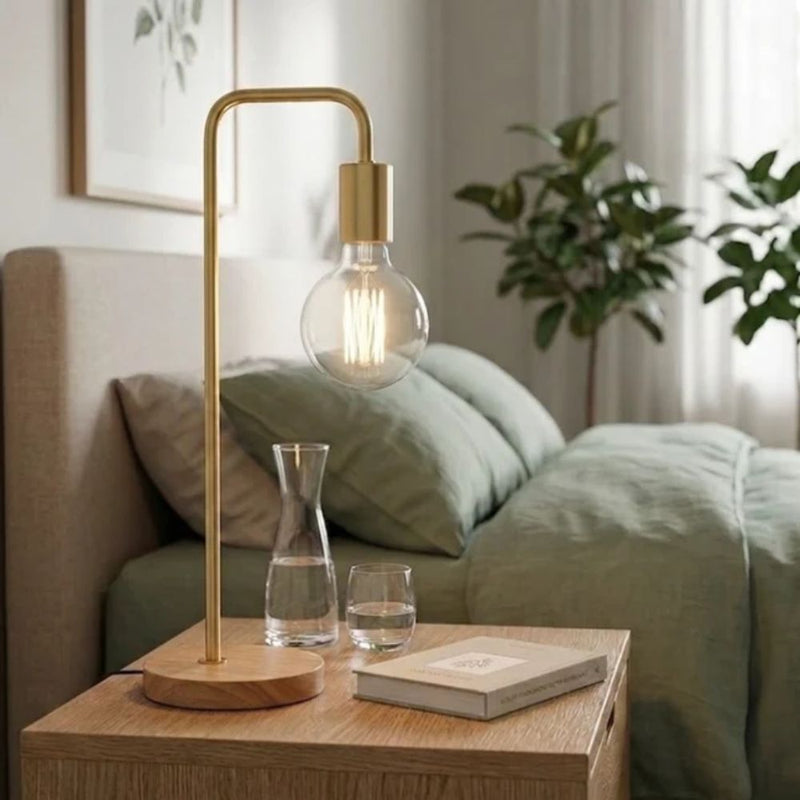 A modern table lamp with a gold-finished metal stand and base, featuring a clear glass globe bulb with a visible filament, placed on a wooden bedside table in a softly lit bedroom - Lighting.co.za