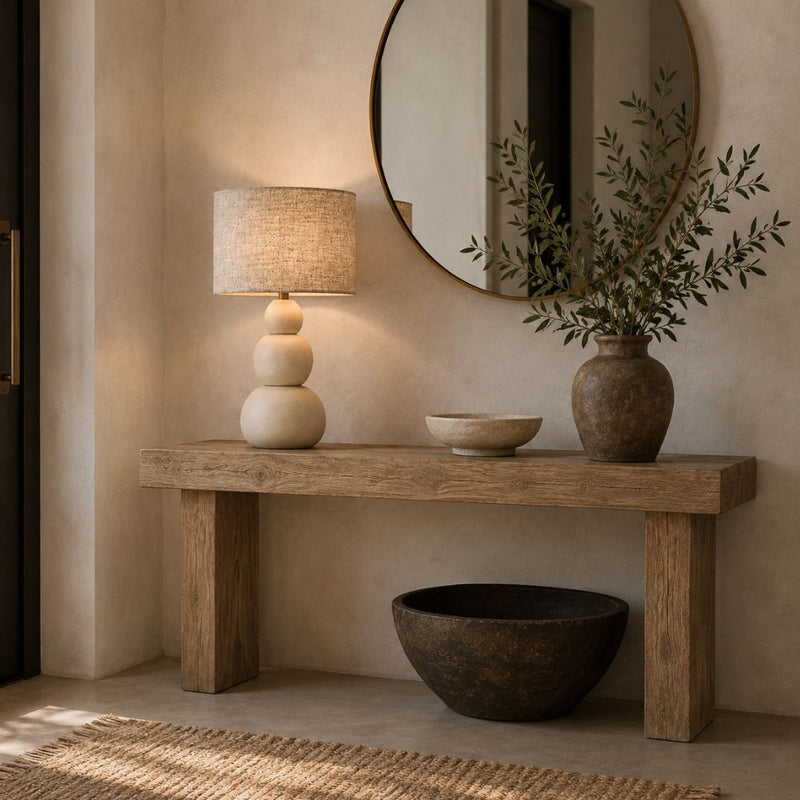 A rustic wooden console table in a warm, neutral-toned entryway, styled with a tiered ceramic table lamp, a small bowl, a large textured vase with greenery, and a dark bowl on the floor - Lighting.co.za