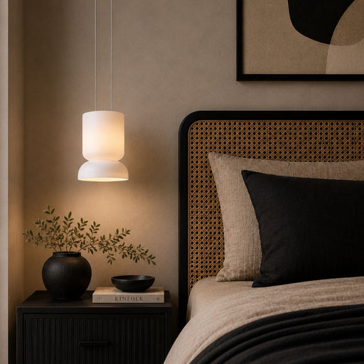 A modern two-tiered pendant light with a glowing white glass cylindrical shade hangs beside a bed with a woven cane headboard, beige bedding, and a black accent pillow in a warm, minimalist bedroom setting - Lighting.co.za