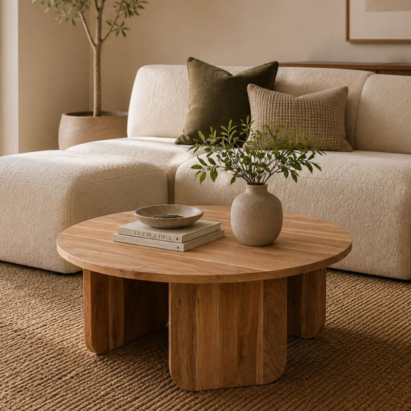 A round wooden coffee table in a warm natural finish, styled in a cozy living room setting with a cream bouclé sofa, decorative pillows, a small vase with greenery, and stacked books on top - Lighting.co.za