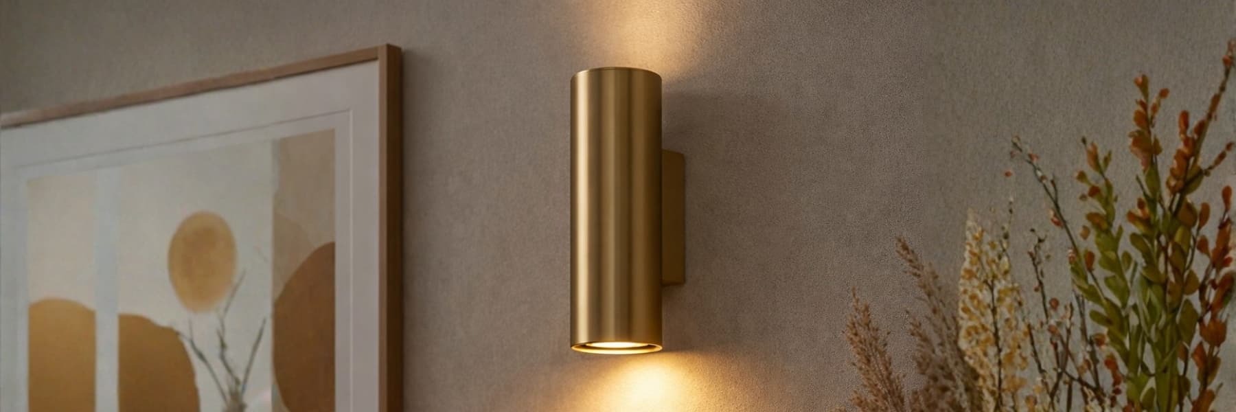 A modern cylindrical wall sconce in a brushed gold finish, mounted on a textured beige wall and casting a warm light downwards and upwards. The sconce is styled in a home interior setting next to a framed abstract print and dried plants.