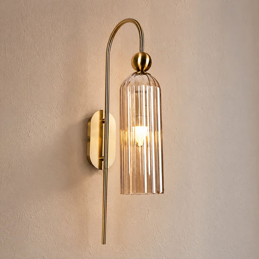 A modern wall-mounted sconce with a curved brass arm, a amber ribbed glass shade, and a warm glowing bulb, mounted on a textured beige wall - Lighting.co.za