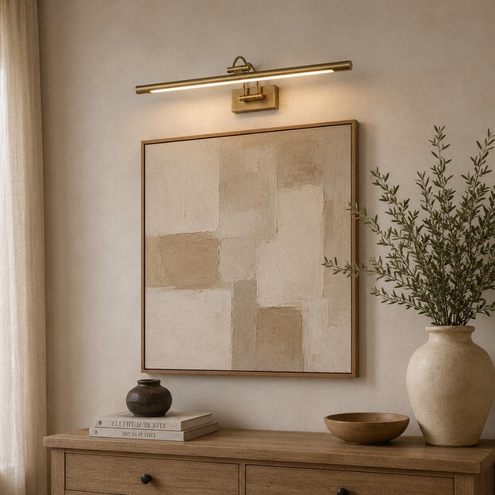 A modern wall-mounted linear light fixture in a warm brass/gold finish, illuminated and mounted above a framed abstract canvas print on a beige wall. Wall Lights Buying Guide. Placement and brightness tips - Lighting.co.za